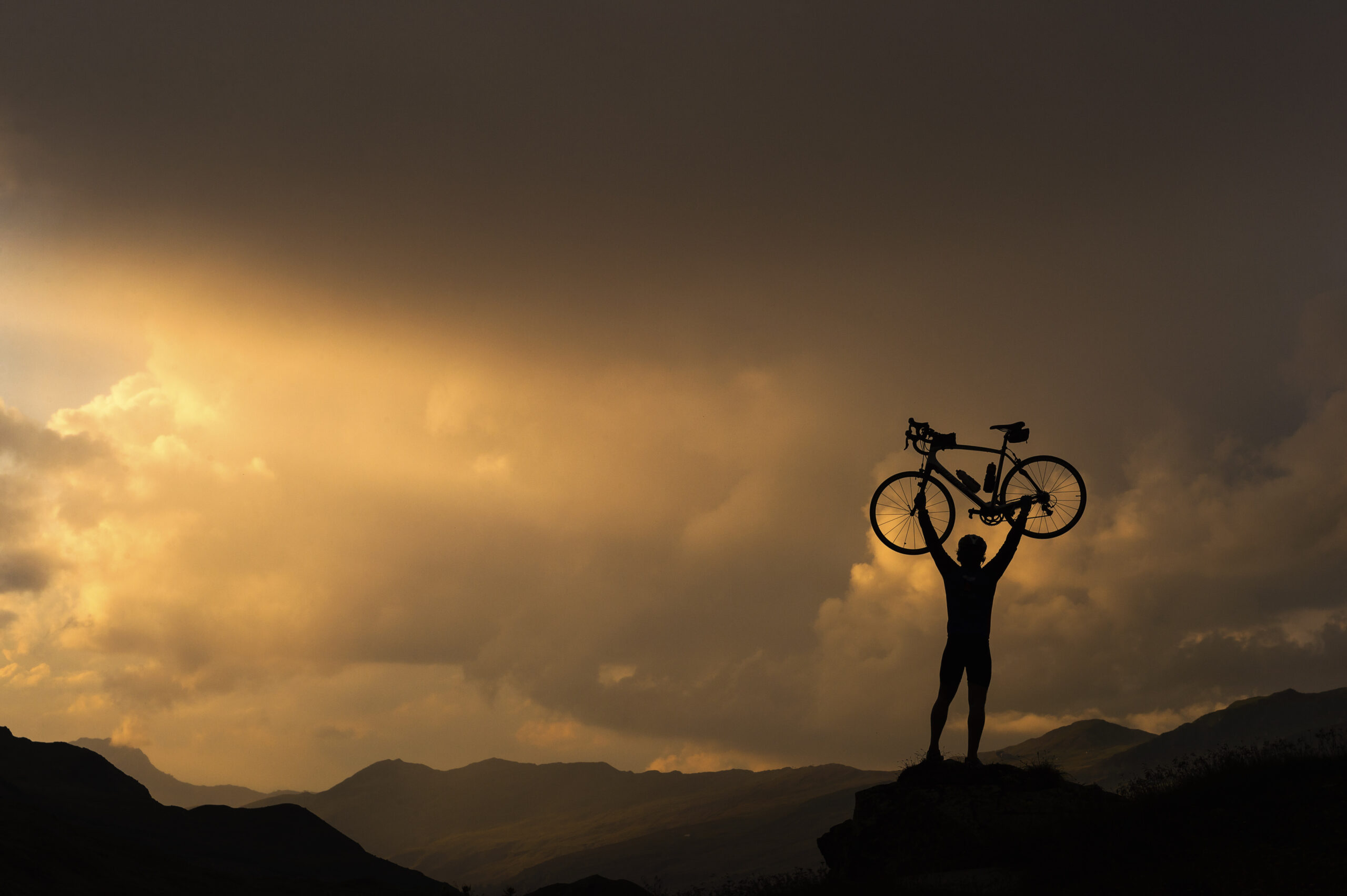 **Alt Text:** "Silhouette of a cyclist triumphantly holding a bike overhead against the backdrop of a vibrant sunset on a mountain landscape, symbolizing achievement, determination, and the spirit of cycling.